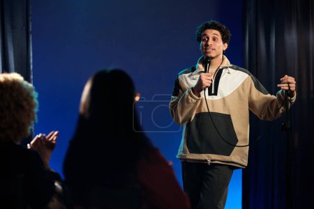 Jeune comédien en tenue décontractée prononçant son monologue au microphone tout en se tenant sur scène et en regardant le public