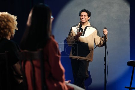 Téléchargez les photos : Heureux jeune humoriste masculin avec microphone prononçant son monologue ou parlant au public tout en se tenant debout sur scène - en image libre de droit
