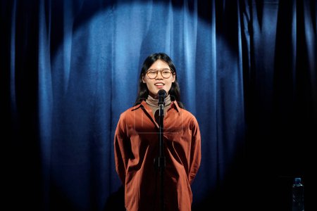 Photo for Young Chinese female comedian in brown shirt standing on stage with blue curtains and speaking in microphone during monologue - Royalty Free Image