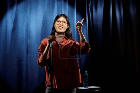 Foto de Mujer asiática joven en gafas y ropa casual de pie en el escenario con cortinas de terciopelo azul y hablando en micrófono - Imagen libre de derechos