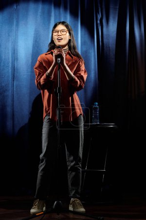Foto de Joven comediante asiática de stand up club de pie en el escenario con cortinas azules y pronunciando monólogo en micrófono - Imagen libre de derechos