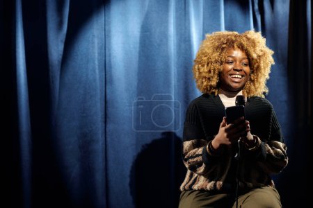 Téléchargez les photos : Jeune femme joyeuse debout humoriste défilant dans le smartphone et parlant au microphone pendant la performance sur scène - en image libre de droit