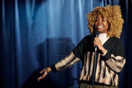 Joven mujer sonriente de pie comediante hablando en micrófono durante la actuación en el escenario, mientras que de pie contra cortinas de terciopelo azul