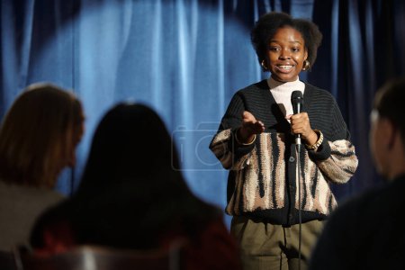 Foto de Joven comediante afroamericana de pie frente a la audiencia en el escenario y hablando en micrófono durante el monólogo - Imagen libre de derechos