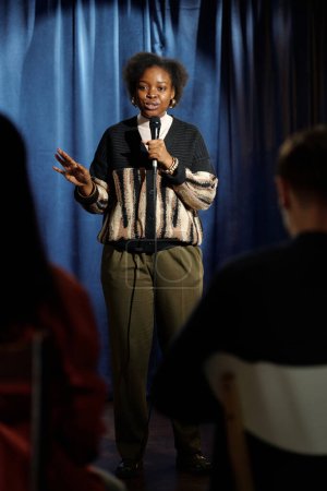 Téléchargez les photos : Jeune présentateur afro-américain en tenue décontractée parlant au microphone tout en se tenant sur scène devant le public pendant le monologue - en image libre de droit