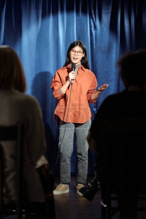 Téléchargez les photos : Jeune comédien de stand up club parlant au microphone debout sur scène avec des rideaux bleus éclairés par un réflecteur devant le public - en image libre de droit