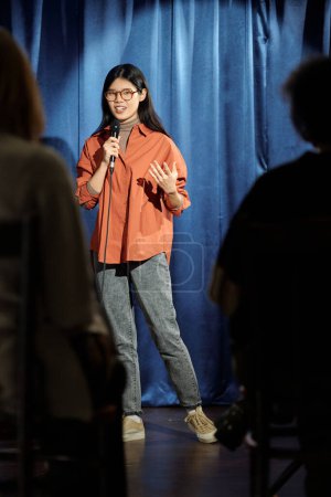 Asian female presenter or participant of stand up show speaking in microphone in front of audience while standing on stage