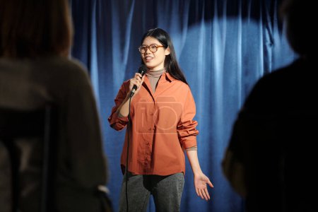 Téléchargez les photos : Jeune comédienne de stand up club debout sur scène avec des rideaux bleus et prononçant monologue devant le public - en image libre de droit