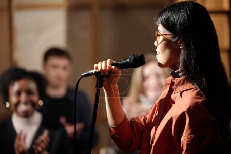 Young female comedian in eyeglasses standing in front of camera against audience and speaking in microphone during performance