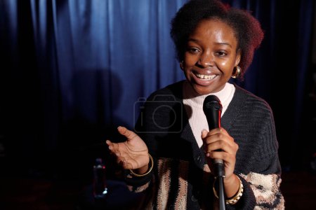 Téléchargez les photos : Joyeux jeune comédienne du club moderne debout tenant le micro tout en parlant devant le public sur scène - en image libre de droit