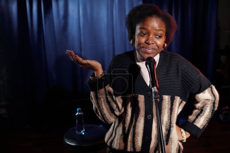Téléchargez les photos : Jeune artiste afro-américaine ou actrice de stand up club parlant au microphone tout en se tenant sur scène avec des rideaux bleus - en image libre de droit