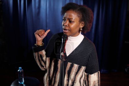 Téléchargez les photos : Jeune femme afro-américaine parlant au microphone et gesticulant debout sur scène contre des rideaux bleus pendant le monologue - en image libre de droit