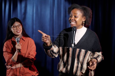 African American woman pronouncing monologue and pointing at audience while standing on stage against Asian girl with microphone