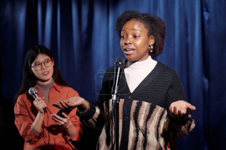 Joven comediante femenina de stand up show pronunciando monólogo mientras está de pie en el escenario frente a la audiencia contra la chica con teléfono inteligente