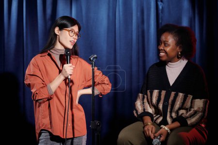 Joven comediante asiática hablando en micrófono y mirando a la artista afroamericana sentada contra cortinas azules en el escenario