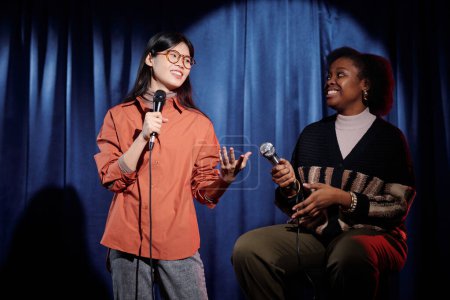 Young Asian woman with microphone pronouncing monologue during stage performance while standing next to another comedian