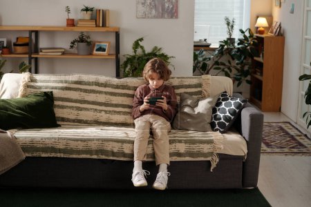 Téléchargez les photos : Mignon petit garçon assis sur un canapé confortable dans un salon spacieux à la maison et regarder la vidéo en ligne sur l'écran du smartphone - en image libre de droit
