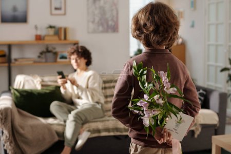 Téléchargez les photos : Vue arrière du garçon tenant un bouquet de fleurs et une carte postale faite à la main pour sa mère tout en se tenant devant une femme avec un smartphone - en image libre de droit