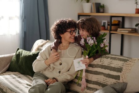 Téléchargez les photos : Garçon mignon avec carte de vœux et bouquet de fleurs embrassant sa mère et la regardant tout en la félicitant pendant les vacances de printemps - en image libre de droit