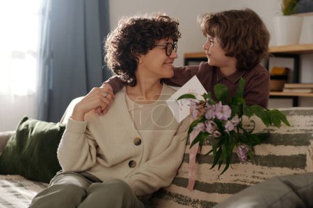 Téléchargez les photos : Garçon affectueux avec un bouquet de fleurs et une carte de vœux regardant sa mère heureuse tout en l'embrassant pendant la célébration à la maison - en image libre de droit