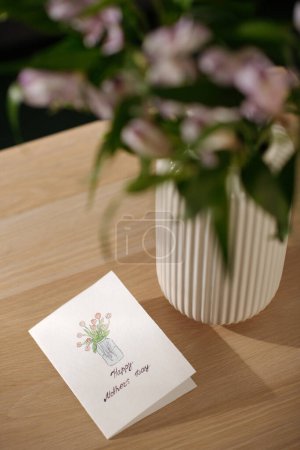Téléchargez les photos : Partie de la table en bois avec carte postale à la main avec salutations pour la fête des mères et dessin de bouquet de tulipes rouges dans un vase - en image libre de droit