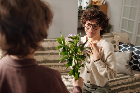 Young surprised woman in eyeglasses looking at bunch of flowers being passed by her little son standing in front of her during congratulations