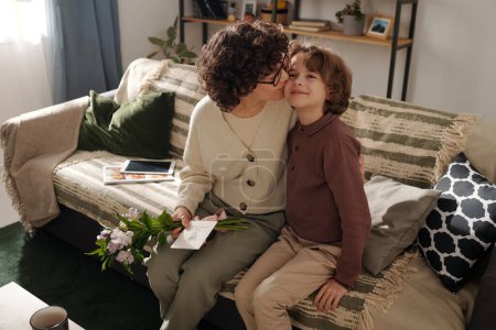 Affectionate mother with bunch of flowers and postcard embracing her adorable son and kissing him on cheek while sitting on couch