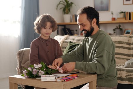 Téléchargez les photos : Petit garçon aidant son père à nouer un bouquet de fleurs pour la mère avec un ruban de soie tout en se tenant près d'une petite table avec du papier et des crayons - en image libre de droit