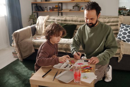 Téléchargez les photos : Adorable garçon peinture à l'aquarelle tout en étant assis à côté de son père avec du crayon lors de la préparation de cartes postales faites à la main pour la fête des mères - en image libre de droit