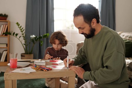 Photo for Bearded man with highlighter bending over small table and drawing something on paper against his son preparing greeting card - Royalty Free Image