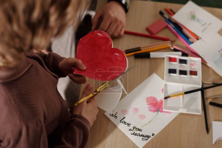 Téléchargez les photos : Angle élevé des mains de l'enfant découpant le coeur de papier rouge sur la table avec des crayons, des cartes postales faites à la main, des aquarelles et des surligneurs - en image libre de droit