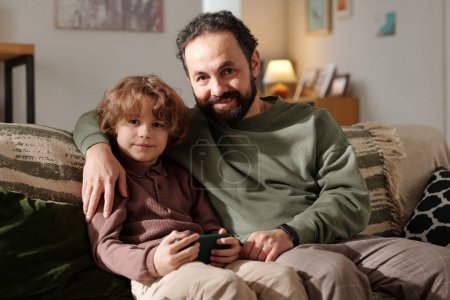 Happy young affectionate man embracing his cute little son with mobile phone while both sitting on couch and looking at camera