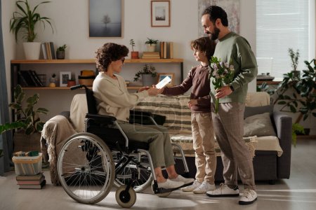 Happy young man and cute boy congratulating their mother and wife in wheelchair on women holiday while standing in front of her