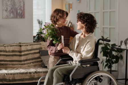 Cute boy holding bouquet of flowers and handmade postcard and looking at his mother in wheelchair while congratulating her on holiday