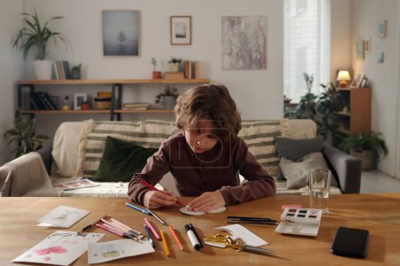 Téléchargez les photos : Garçon diligent avec crayon rouge créant une carte postale faite à la main pour sa mère tout en étant assis à table avec des papiers, aquarelles et surligneurs - en image libre de droit