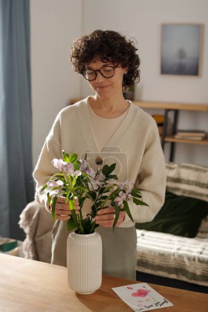 Téléchargez les photos : Jeune femme brune mettant un bouquet de fleurs dans un vase tout en se tenant à table dans l'environnement de la maison et se préparant pour les vacances de printemps - en image libre de droit