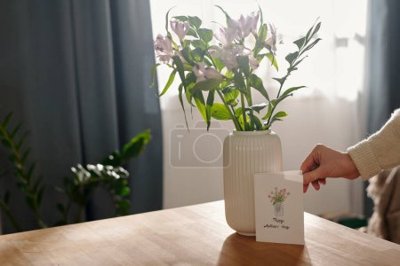Téléchargez les photos : Main de jeune femme méconnaissable mettant carte postale à la main pour la fête des mères à côté du vase blanc avec un bouquet de fleurs - en image libre de droit