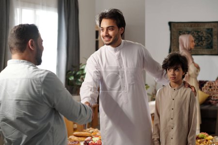 Foto de Mediana toma de joven musulmán y su hijo vistiendo thobes saludo pariente masculino en Eid al-Fitr - Imagen libre de derechos