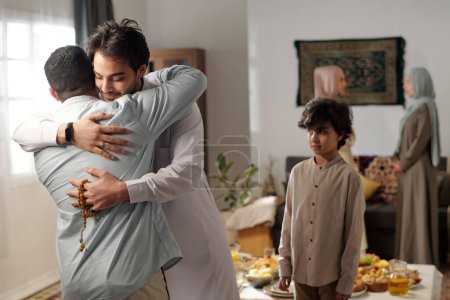 Medium shot of young Muslim man wearing thobe greeting male relative with hug on Eid al-Fitr, his son standing next to him