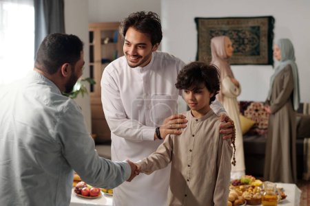 Foto de Mediana toma de hombre musulmán moderno y su hijo vistiendo thobes saludo pariente masculino en Eid al-Fitr, dos mujeres hablando en segundo plano - Imagen libre de derechos