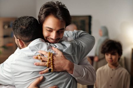Medium closeup of young Middle Eastern man greeting his friend or relative with hug while gathering with family on Islamic holiday