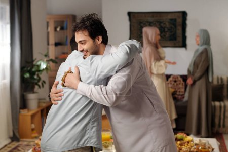 Foto de Vista lateral media de dos hombres musulmanes saludándose con abrazo en Eid Al-Fitr, dos mujeres con hiyabs hablando en segundo plano - Imagen libre de derechos