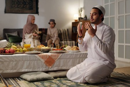 Téléchargez les photos : Mise au point sélective du jeune homme musulman assis sur le sol par table festive priant salah, sa femme et son fils lisant le Coran en arrière-plan - en image libre de droit
