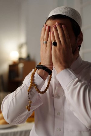 Vertical medium closeup of unrecognizable young Muslim man wearing white clothes with taqiyah covering face with hands while praying salat