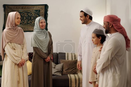 Téléchargez les photos : Hommes, femmes et enfants musulmans modernes portant des vêtements modestes debout dans le salon et discutant lors d'une réunion de famille sur l'Aïd Al-Fitr - en image libre de droit
