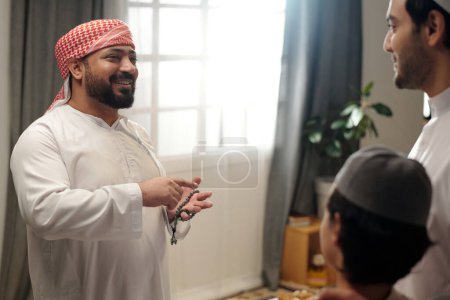 Foto de Hombre musulmán alegre sosteniendo cuentas de oración charlando con su hermano menor y sobrino en la reunión familiar en Eid Al-Fitr - Imagen libre de derechos