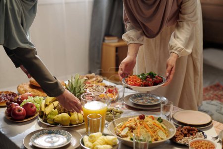 Two unrecognizable Muslim women wearing plain dresses and hijabs setting festive table for Eid Al-Fitr celebration with delicious dishes