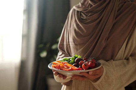 Téléchargez les photos : Gros plan moyen de femme musulmane méconnaissable portant du hijab tenant une assiette avec des tomates cerises, de la laitue et des tranches de poivron dessus - en image libre de droit