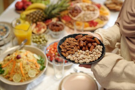 High angle view closeup of hands of unrecognizable Muslim woman holding plate full of almonds, dates and peanuts while setting festive table