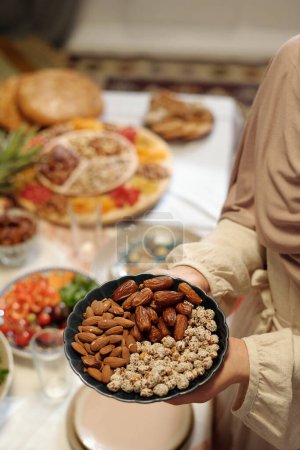 Großaufnahme der Hände einer nicht wiederzuerkennenden muslimischen Frau, die einen Teller voller süßer Mandelnüsse, Datteln und Erdnüsse hält und den festlichen Tisch für Uraza Bayram deckt